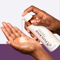 A person with a medium-to-deep skin tone pumps a creamy product from a white and gold Flourish Nourishing Cream-Oil bottle into their palm. 