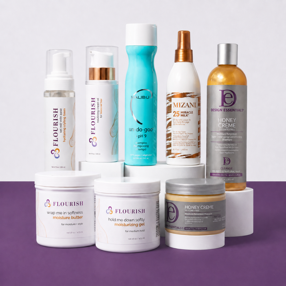 A collection of hair care products from the brands Flourish, Malibu, Design Essentials, and Mizani, arranged on white display blocks against a split white and light gray background with a purple surface. The set includes various bottles, jars, and a black hairbrush.